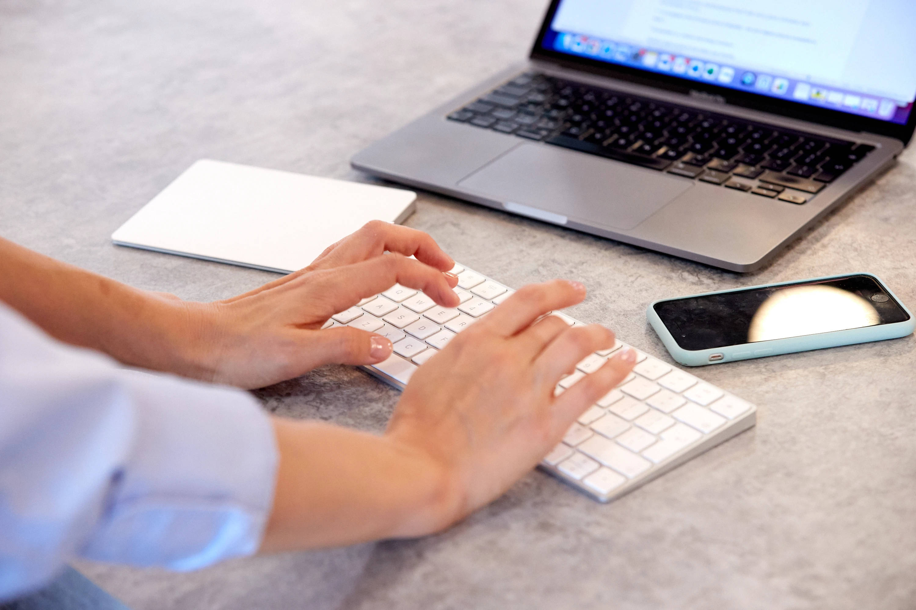 A photo of woman's hands typing on a keyboard. A laptop and a mobile phone are in the background.