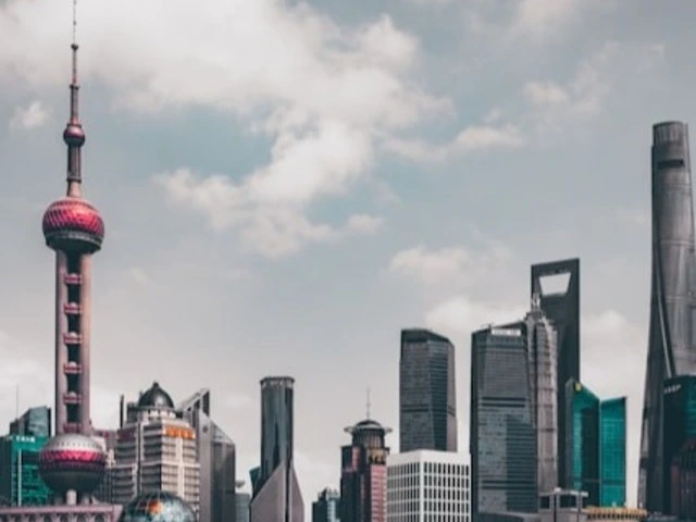 Modern city skyline of Beijing, China, featuring a mix of iconic skyscrapers and unique architectural structures. Prominently on the left is a tall tower with a spherical observation deck, the Oriental Pearl Tower. The surrounding buildings vary in design, with some featuring sharp, angular forms and others with sleek, curved surfaces. The sky above is cloudy.