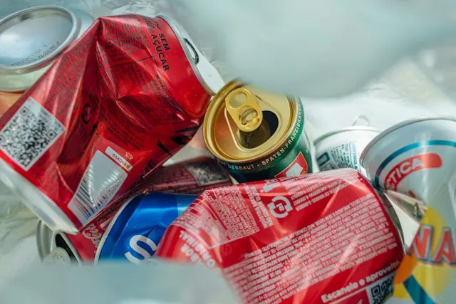 Collection of crumpled and discarded aluminum cans inside a trash bag or recycling bin. The cans are of various colors, including red, blue, and green, with some still partially intact and others crushed. The pull-tabs on some cans are visible, with one can's top prominently showing its golden pull-tab. The background is slightly blurred.