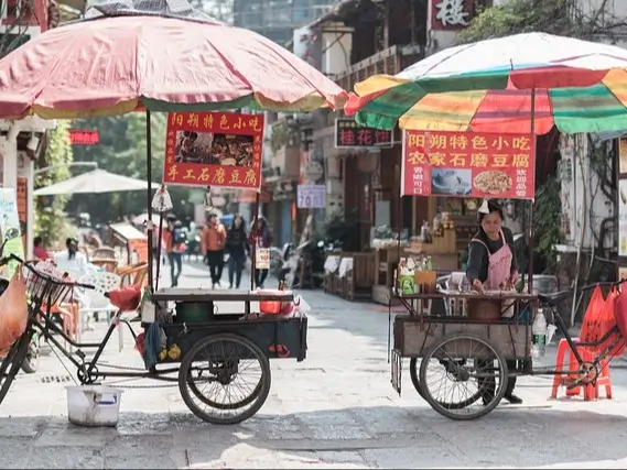 Two street food carts positioned side by side in an outdoor market setting in China. Each cart has a large umbrella providing shade. The left cart has a red umbrella, and the right cart has a multicolored umbrella. A vendor stands behind the right cart, wearing an apron. The scene is set on a street with other shops and people in the background, creating a lively atmosphere. Bright signs in Chinese characters are displayed above both carts.