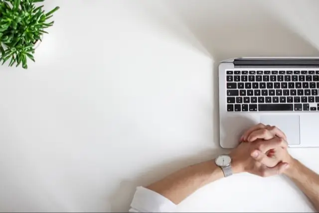 Minimalist workspace with a white background. In the top right corner, a silver laptop with a black keyboard is partially visible. Below the laptop, a pair of hands are resting on the desk, fingers interlocked, with the person wearing a wristwatch. On the top left corner, there is a small green plant in a white pot, adding a touch of nature to the clean, organized setup.