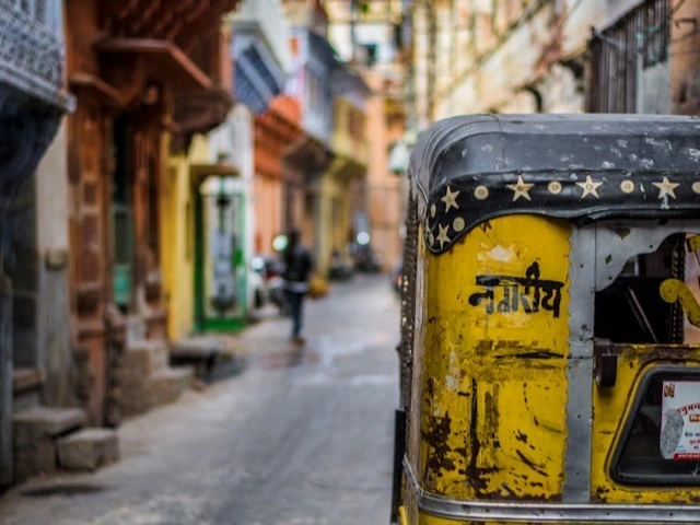 Rear view of a yellow auto-rickshaw in a narrow, colorful street in Jodhpur, India. The rickshaw is weathered, with chipped paint and some dirt, and has text in a local script on its back. The street is lined with closely packed buildings, featuring various architectural styles and colors. In the background, a person is walking away.