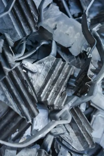 Close-up of a pile of crumpled and twisted magnesium sheets. The sheets are dented and bent, with some pieces having ridged or corrugated surfaces. The metal has a dull, matte finish, with varying shades of gray and silver.