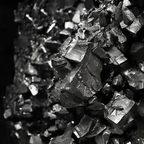 Close-up view of a cluster of dark, shiny mineral crystals made of zinc. The crystals have sharp, angular edges and exhibit a reflective, almost mirror-like surface, with a deep black or dark gray coloration. The texture is rugged and irregular, with light glinting off the facets, highlighting the geometric shapes of the crystals.