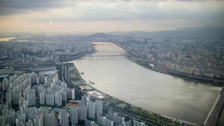 The image shows an expansive aerial view of a modern cityscape in South Korea dominated by a wide river running through the center. The city is densely packed with high-rise buildings, particularly clustered near the riverbanks. Multiple bridges span the river, connecting the two sides of the city. The sky is overcast, casting a muted, grayish light over the scene, giving it a calm, subdued atmosphere. 