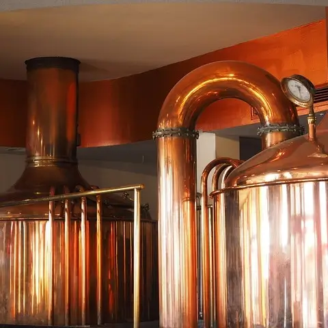 Shiny copper brewing equipment inside a brewery. Prominent features include large, cylindrical vats and tanks connected by thick copper pipes. The surfaces of the equipment are polished and reflect the warm light, giving the scene a rich, golden glow. The brewing tanks are capped with domed tops, and a gauge is visible on one of the pipes, indicating its industrial use.