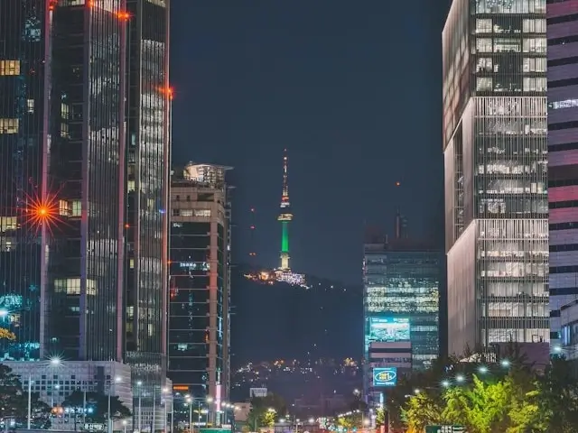 Nighttime cityscape in Seoul, South Korea, with tall, modern skyscrapers lining both sides of a broad avenue. In the center background, an observation tower is illuminated in green and stands on a hill overlooking the city. The buildings have glowing lights in their windows, and the street below is dotted with moving cars and streetlights.