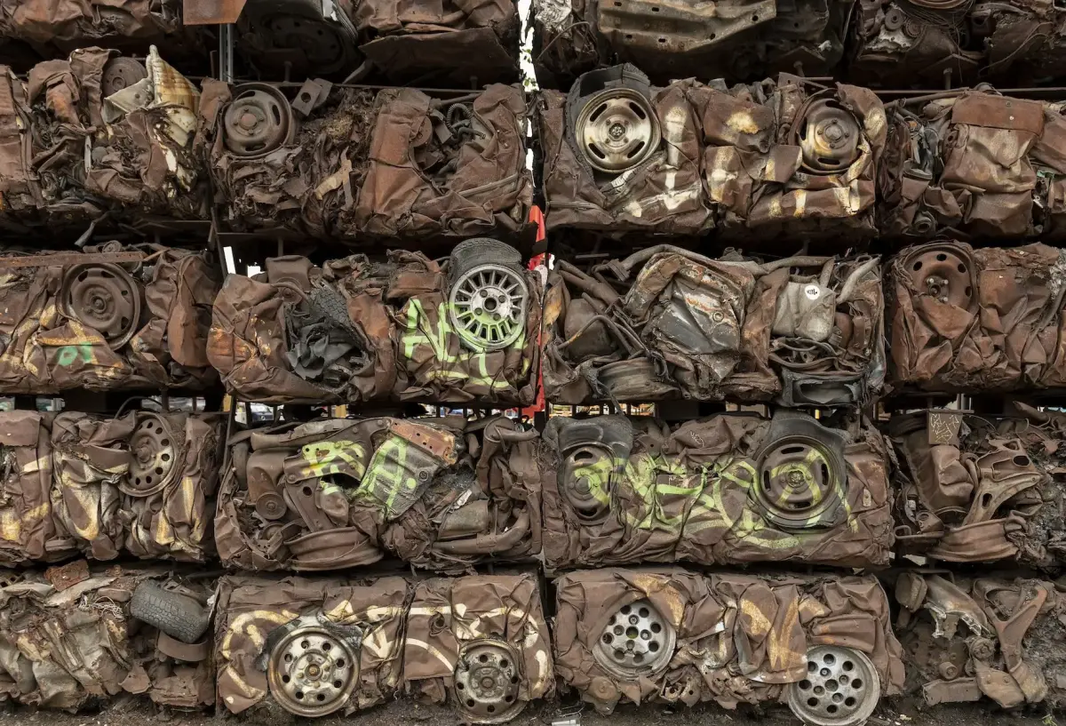 Large stack of crushed cars, compacted into rectangular blocks. The cars are tightly compressed, with various parts like wheels, tires, and twisted metal frames visible within the densely packed mass. The colors are mostly rusted brown, with some faded paint and graffiti on the crushed surfaces. The stack forms a grid-like pattern, emphasizing the industrial process of recycling or scrapping vehicles.