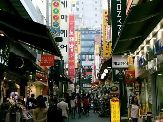 Bustling, narrow street in a South Korean city, lined with shops and signs in various languages, predominantly Korean. The street is crowded with people walking in both directions. Bright, vertical signs in different colors hang from the buildings, advertising various businesses. The shops on either side display products, with some items extending onto the sidewalk.