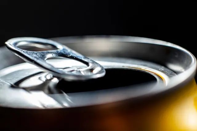 Close-up of the top of an opened aluminum can, focusing on the pull-tab and the rim of the can. The pull-tab is lifted, revealing the open drinking hole. The metal surface of the can is shiny and reflective, with subtle highlights and shadows that emphasize its smooth texture. The background is dark, making the metallic sheen of the can stand out.