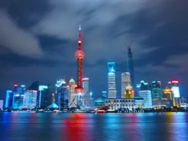 Stunning night view of a Shanghai along a waterfront. The skyline is dominated by the Oriental Pearl Tower, illuminated in bright red, with its distinctive spherical structures. Surrounding skyscrapers are lit up in various colors, reflecting in the calm waters below. The sky is partly cloudy.