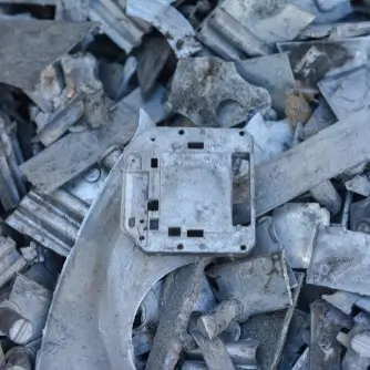 Pile of discarded magnesium parts, including pieces of sheet metal, broken machinery components, and various other industrial scraps. In the center, there is a square, hollow metal piece with multiple cutouts, a frame and casing from a device. The metal is worn, tarnished, and shows signs of corrosion.