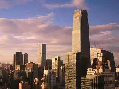 Beijing city skyline during sunset, featuring tall skyscrapers with varying architectural styles. The tallest building dominates the scene, with a sleek, reflective surface that catches the warm light of the setting sun. The sky is filled with soft, pink and purple clouds, creating a serene backdrop.