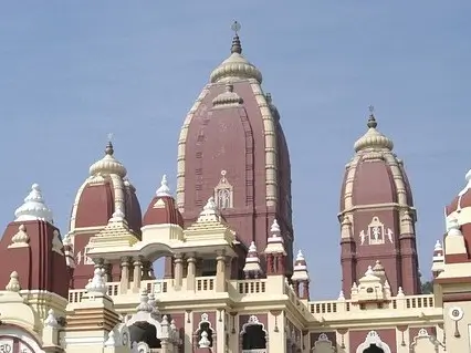 Ornate domes and towers of a Hindu temple in India against a clear blue sky. The temple's architecture features tall, reddish-brown domes with intricate white detailing, including carvings and decorative elements. The central dome is the tallest, flanked by smaller domes of similar design.