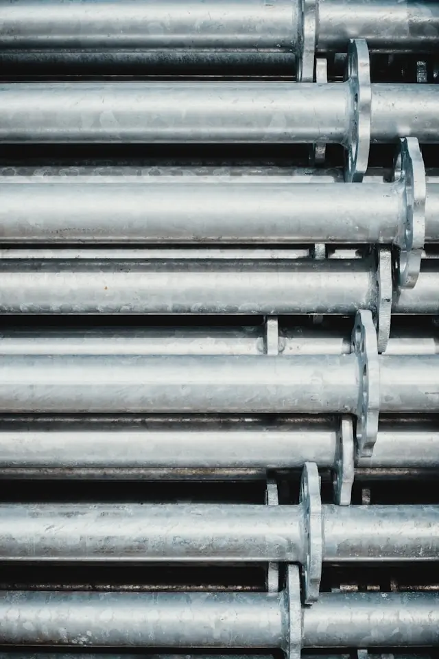 Stack of large, aluminum pipes arranged horizontally in a neat, parallel formation. The pipes are uniform in size, with smooth, shiny surfaces that reflect light. Each pipe has a flange at regular intervals, likely for connecting to other pipes or structures. 