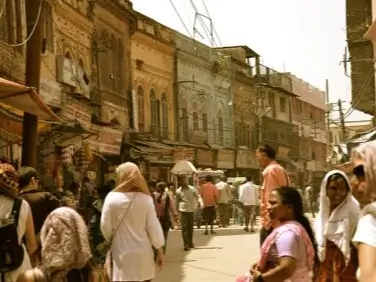 Bbusy street scene in a market area in India. The street is lined with traditional buildings featuring ornate facades in earthy tones of yellow, brown, and blue. People dressed in a variety of traditional and modern attire walk along the street, some engaging in conversation or shopping. 