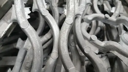 Close-up view of a collection of metallic magnesium components, likely industrial parts or machine pieces. These parts appear to be curved metal strips with circular connectors or joints at various points, giving them a somewhat serpentine or twisted appearance. The material seems to be a dull gray metal, with a slightly rough or unfinished texture. The components are intertwined or stacked together, creating a dense and complex pattern of overlapping shapes.