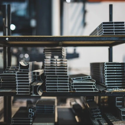 Storage rack filled with neatly stacked aluminum profiles in a workshop or industrial setting. The metal pieces are of various shapes and sizes, including rectangular tubes, square bars, and L-shaped angles. The metal is clean and reflective, indicating it is likely new or unused. The background is blurred, focusing attention on the organized metal inventory.