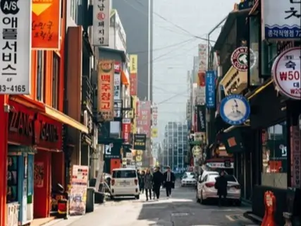 Busy urban street in Seoul, South Korea, lined with buildings, shops, and numerous signs in a mix of Korean and English. The street is narrow, with cars parked along the sides and a few pedestrians walking down the middle. The buildings are closely packed, with colorful storefronts and a variety of advertisements and banners hanging above.