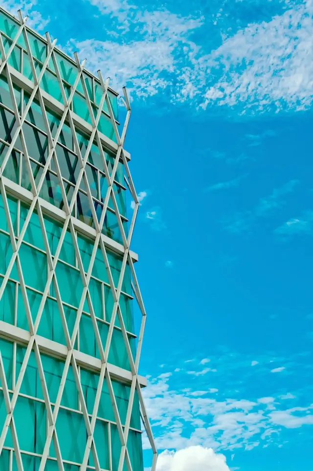 Modern building with a unique architectural design. The building's facade is made of large green-tinted glass panels, crisscrossed by an external metal framework that forms a diamond-shaped pattern. The structure occupies the left side of the image, while the right side is dominated by a bright blue sky with scattered white clouds. The contrast between the building's geometric design and the natural sky creates a striking visual effect.