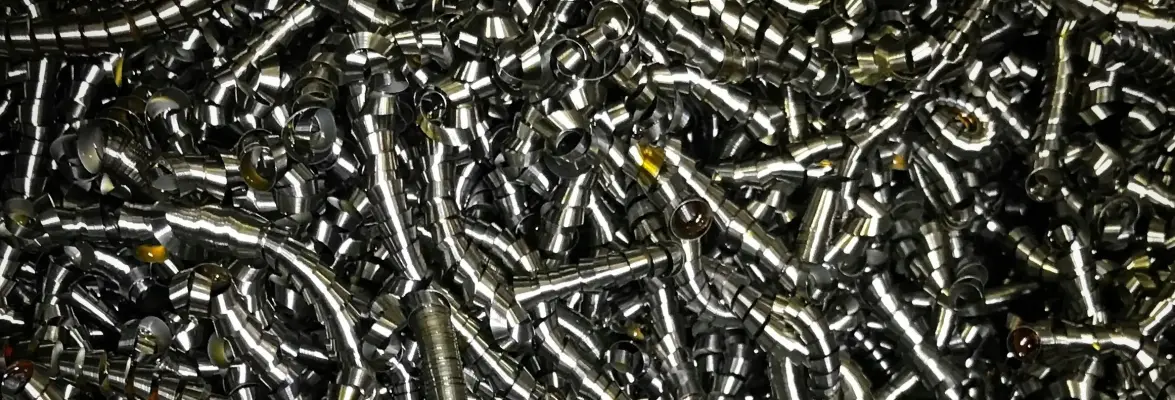 Dense pile of shiny, metallic screws and fasteners. The screws are tightly packed together, creating a textured surface with a uniform metallic sheen. The screws vary slightly in size and shape but are predominantly cylindrical with threads, reflecting light off their smooth surfaces. A few screws have hints of yellow markings or corrosion.