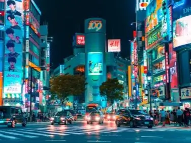 Vibrant, bustling city street at night in Tokyo. The scene is illuminated by numerous bright, colorful neon signs and billboards on both sides of the street, advertising various products and brands. The street is busy with cars and pedestrians, creating a lively atmosphere. The lights from the signs reflect off the wet pavement, enhancing the colorful glow.