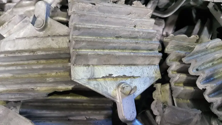 Close-up of a pile of old, weathered metal radiator fins and heat sink components made of magnesium. The metal surfaces are worn and discolored, with a buildup of grime and oxidation, giving them a dull, aged appearance. The fins are stacked haphazardly, with some showing signs of damage or bending.