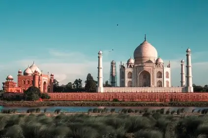 Taj Mahal in Agra, India, bathed in soft daylight. The majestic white marble mausoleum, with its large dome and four minarets, stands prominently against a clear blue sky. To the left, a red sandstone mosque complements the Taj Mahal's grandeur. The foreground features lush green vegetation and a calm river.