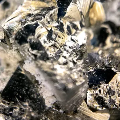 Close-up of a cluster of metallic crystals, likely a mineral or metal ore. The crystals are sharp, with reflective surfaces that catch the light, displaying a mix of shiny silver, black, and gray tones. The intricate details of the crystal structure are visible, with jagged edges and a textured surface. The overall appearance is rugged and raw, highlighting the natural beauty of the mineral in its unrefined state.