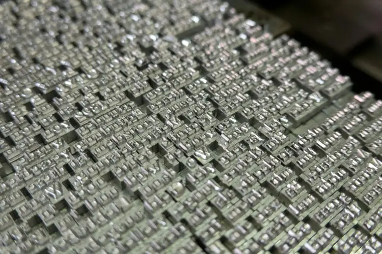 Close-up of a large number of lead type blocks, typically used in traditional printing presses. Each block has a raised letter or character on its surface, arranged in a grid-like formation. The metallic type blocks have a shiny, silver appearance and are densely packed together, creating a textured surface with the distinct shapes of letters and symbols visible. The arrangement suggests these are ready for use in typesetting, where they would be inked and pressed onto paper to produce text.