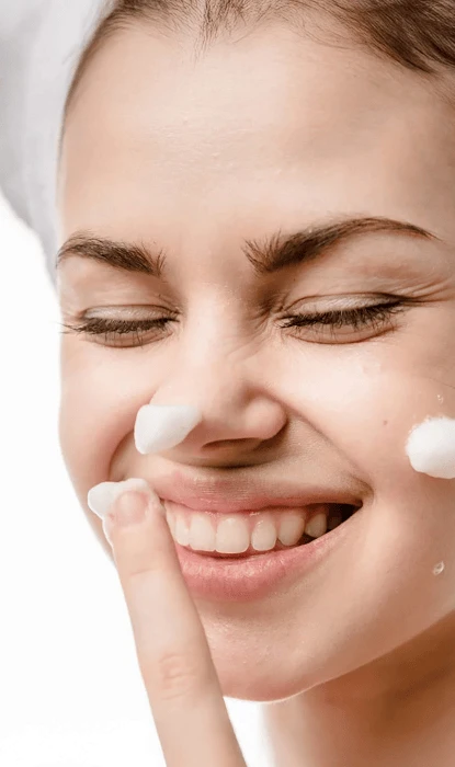 Mujer joven sonriente que lleva una toalla en la cabeza y se aplica espuma de afeitar en la nariz y las mejillas