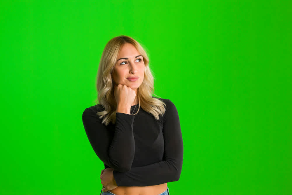 Doubtful pensive woman with thoughtful expression, presses lips together, keeps hand under chin, plans something or makes choice standing in front of a green screen