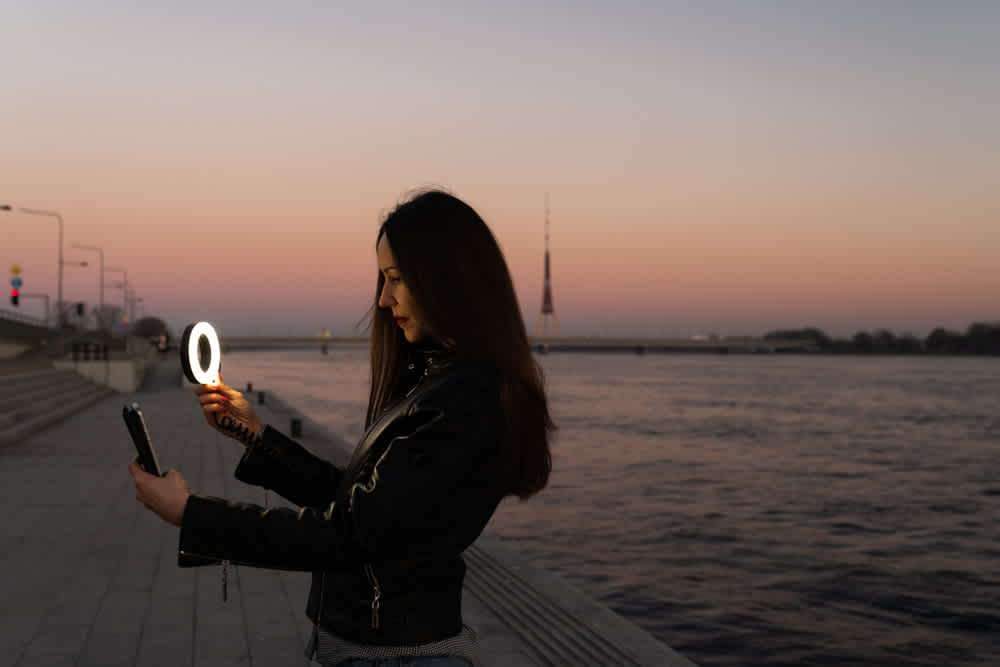 Young woman taking a selfie using a ring flash as a fill light at a sunset with a view over river - selfie video tips