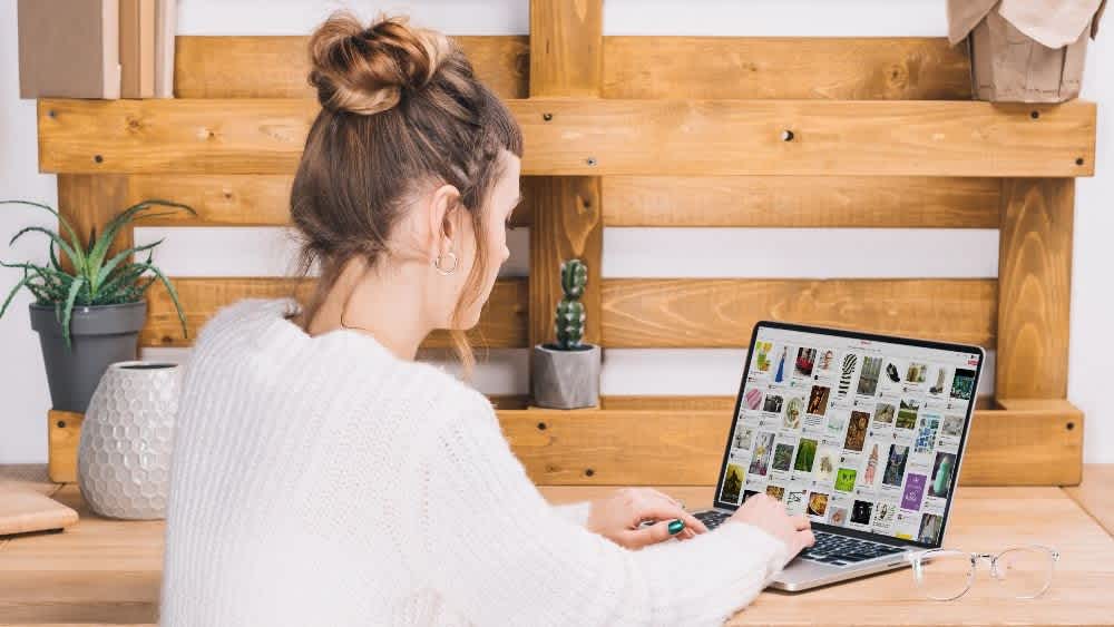 A woman sits at a desk browsing Pinterest on her laptop.