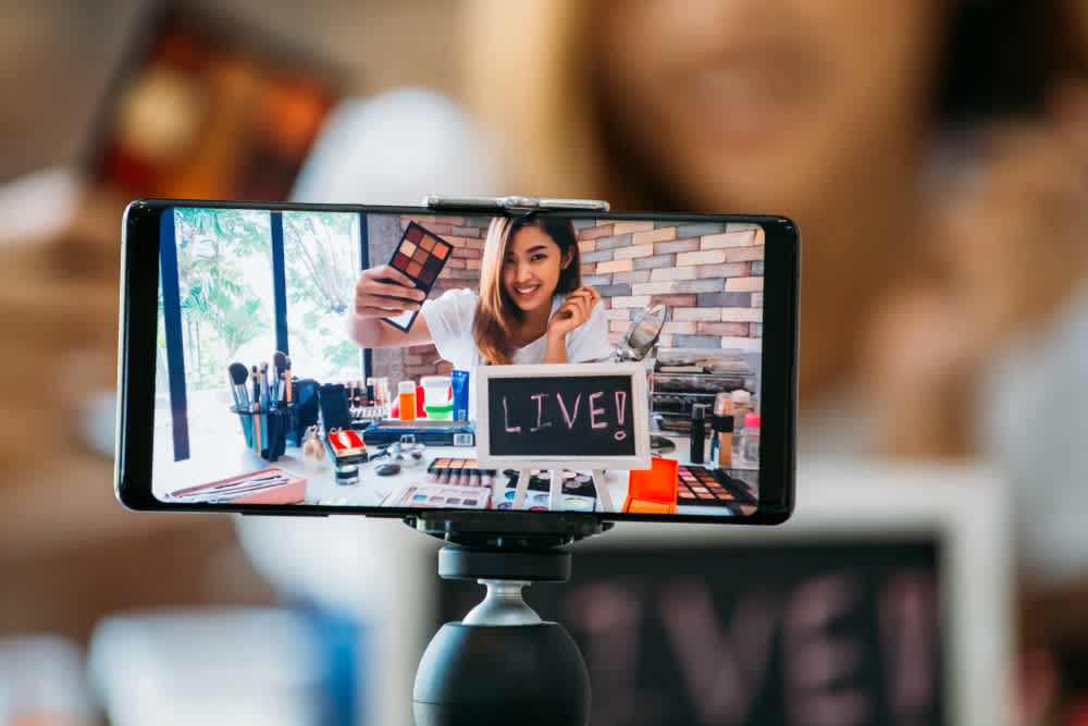 Happy young attractive Asian ethnic model filming vlog on mobile phone on tripod and showing eyeshadows at table with cosmetics