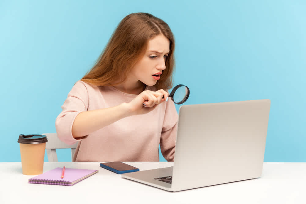 woman journalist looking through magnifying glass with surprised expression at laptop - Fraud in Video Viewing What You Should Know - Clipchamp Blog