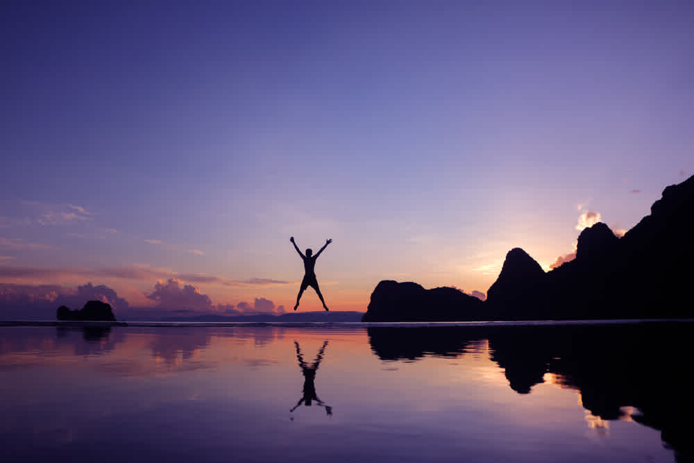 Man having fun jumping in the morning at sunrise on the beach reflections on the water surface sea and mountain background long shot