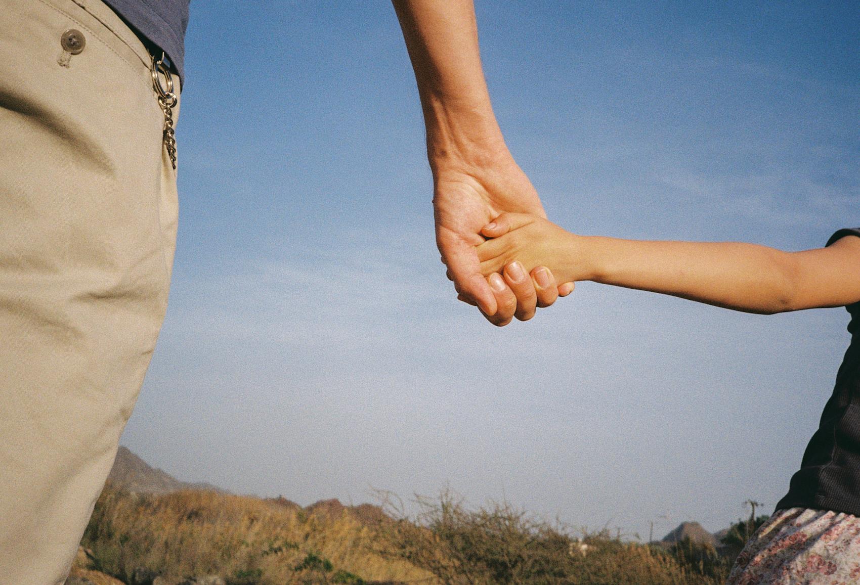 Bildet viser en voksen person og et barn holde hender, med blå himmel i bakgrunnen.