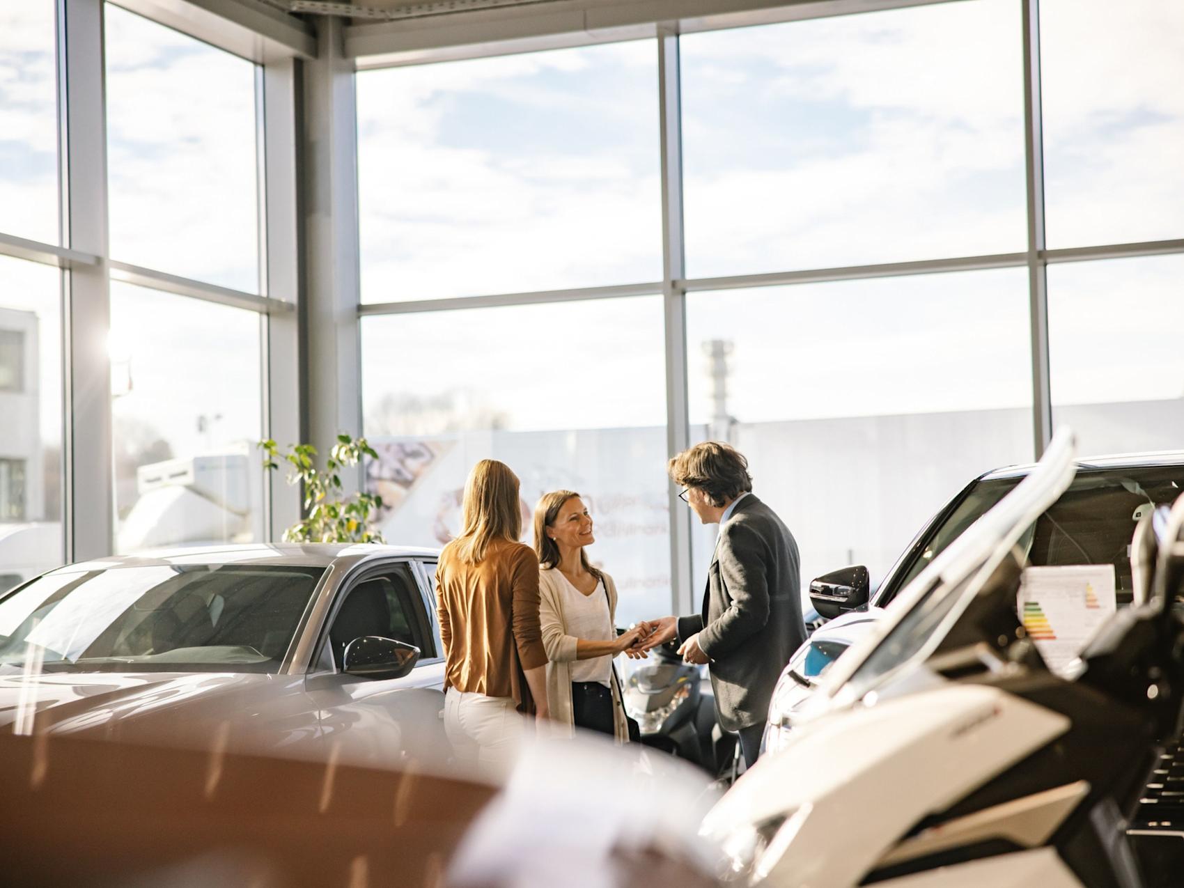 Tre personer i samtale i en lys bilforretning med store vinduer og utstilte biler. Sollys strømmer inn gjennom glasset.