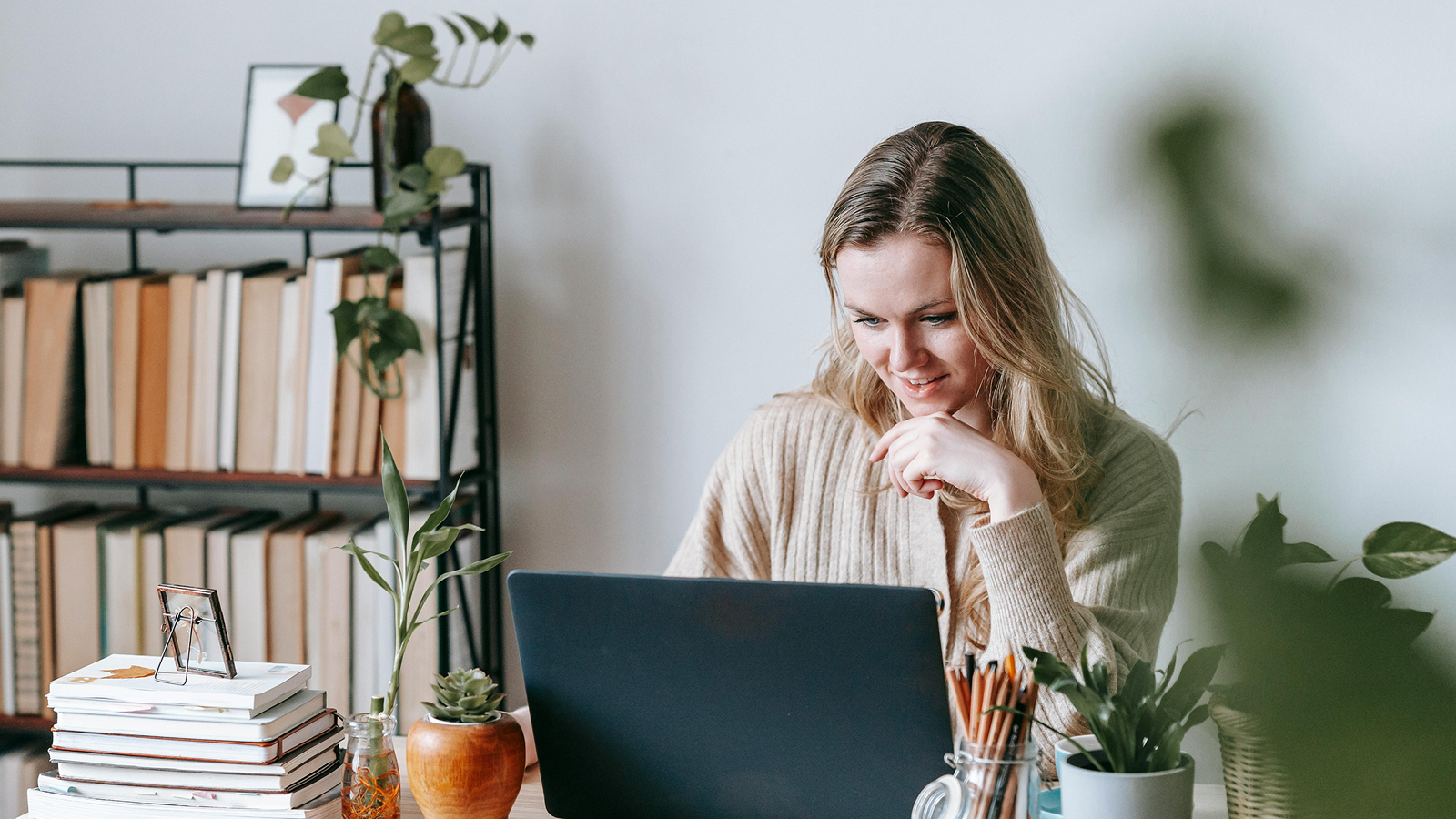Kvinne i beige genser smiler mens hun arbeider på laptop ved et bord med bøker og planter, bokhylle i bakgrunnen.