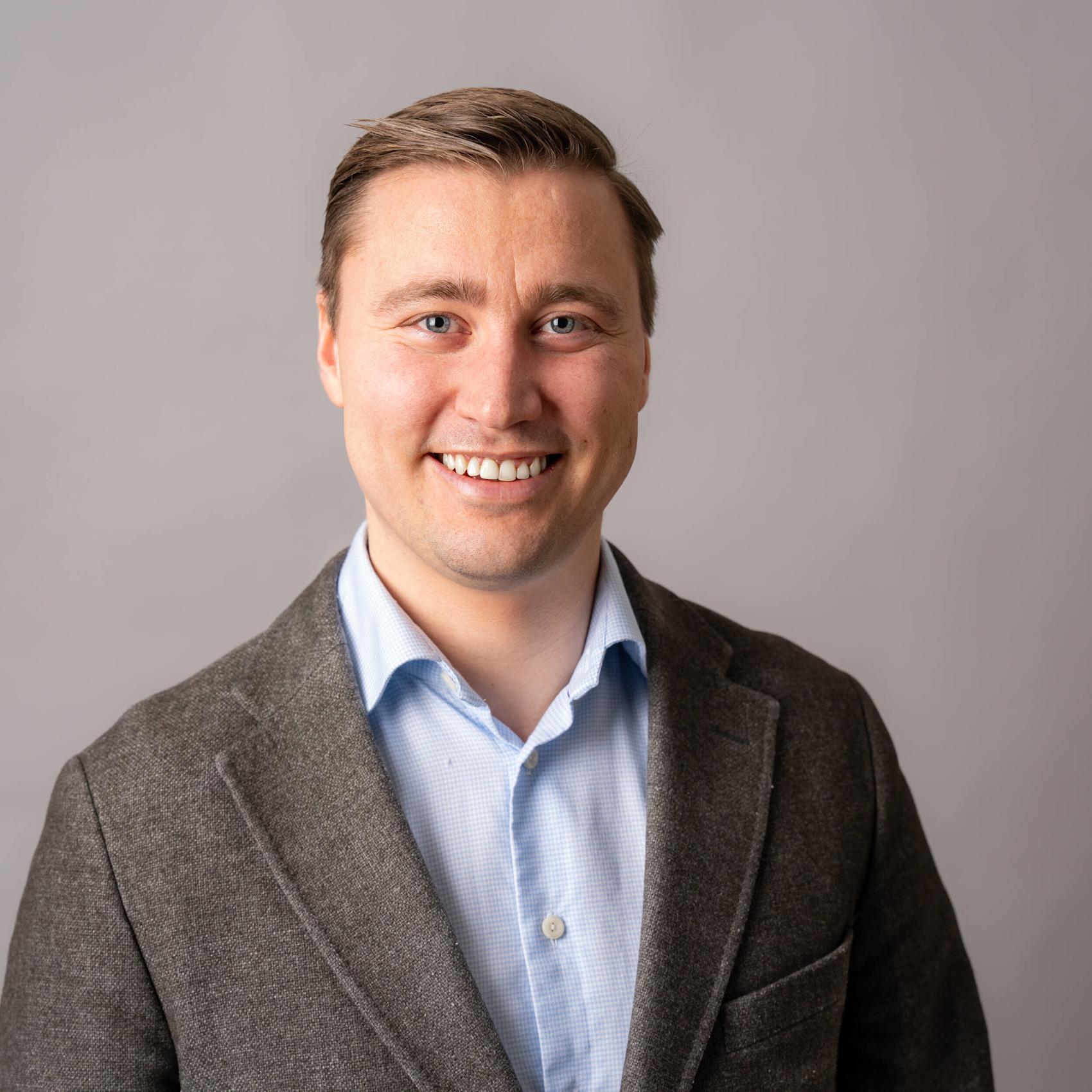 Portrett av en smilende person med blå øyne, kledd i grå dressjakke og lyseblå skjorte mot en nøytral bakgrunn.