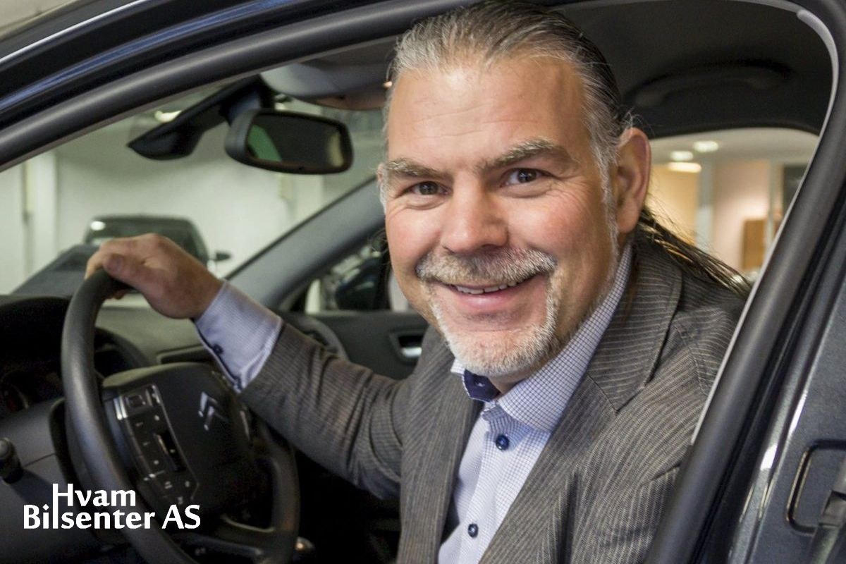 En smilende person i dress sitter bak rattet i en bil i et bilsenter. Hvam Bilsenter AS-logo vises i hjørnet.