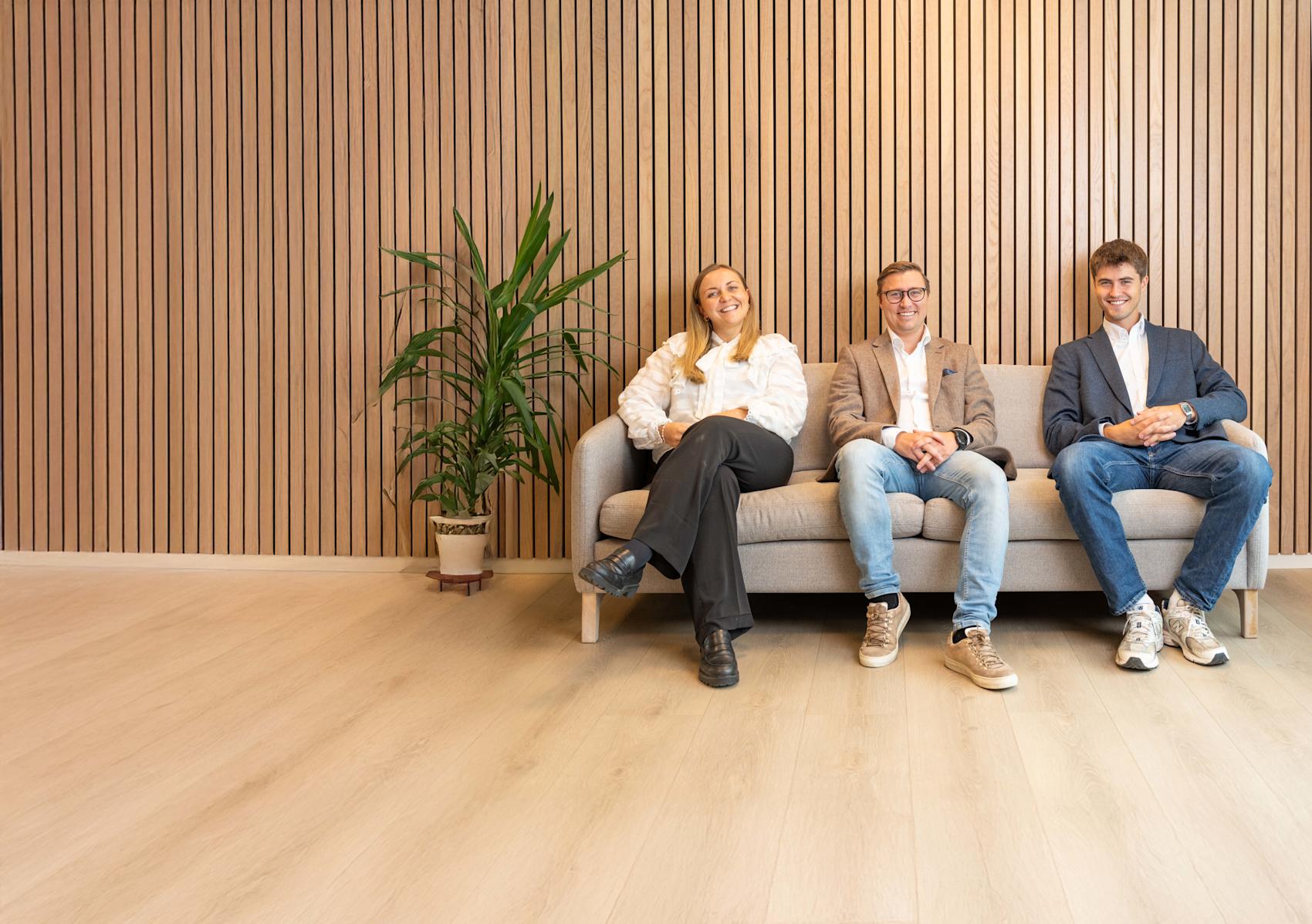 Tre smilende personer sitter på en grå sofa foran en trevegg. En grønn plante er synlig til venstre.