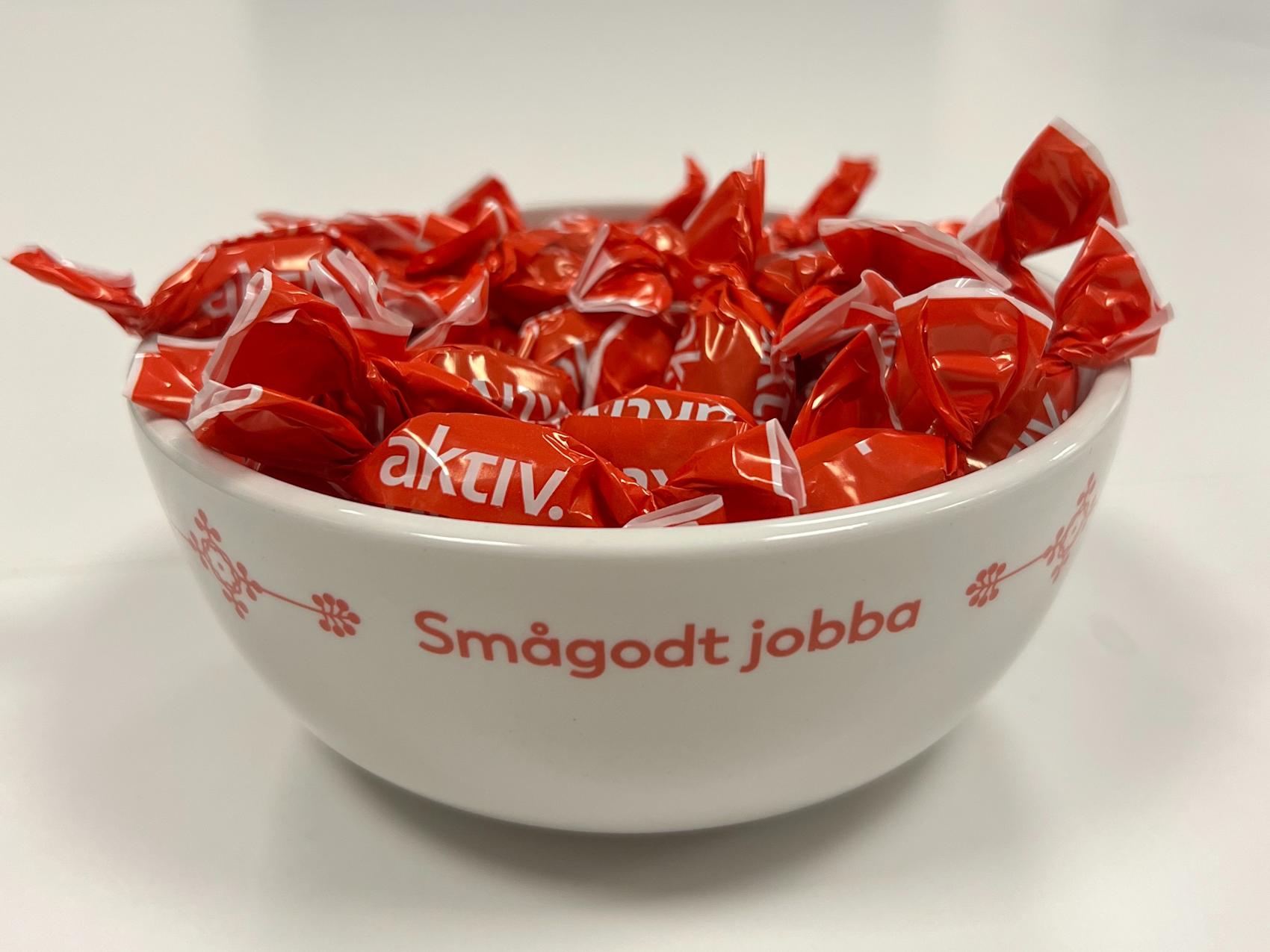 Hvit skål med teksten "Smågodt jobba" fylt med røde Aktiv-drops på et hvitt bord.
