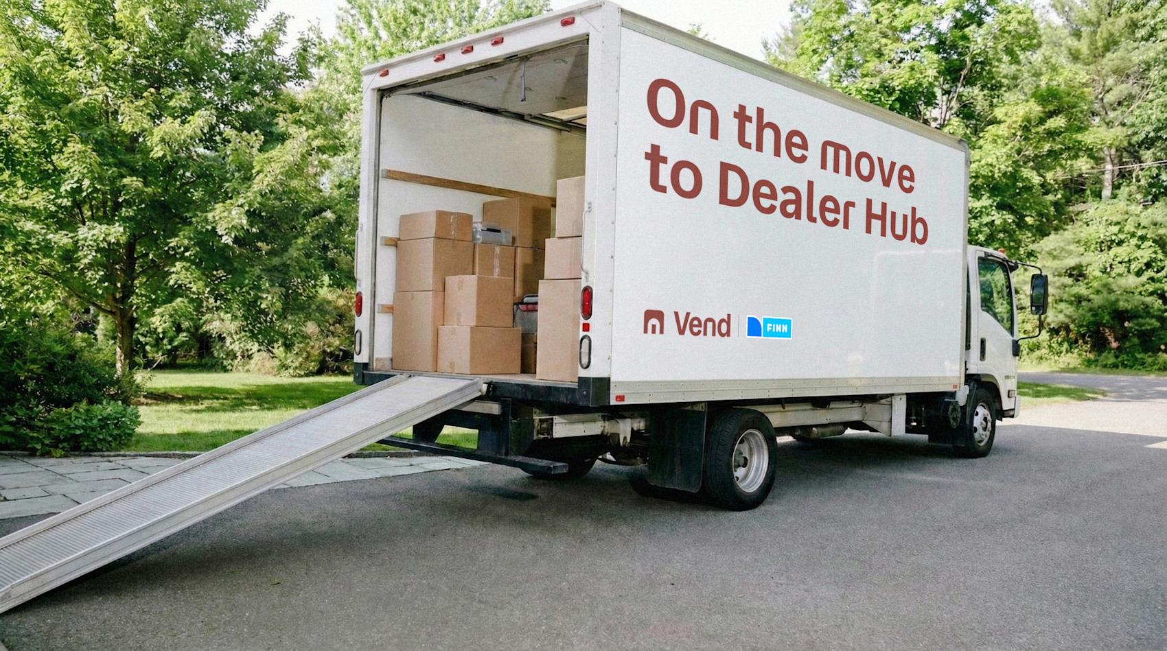Hvit flyttebil med teksten "On the move to Dealer Hub" lastet med esker, parkert i grønt område med rampe ute.