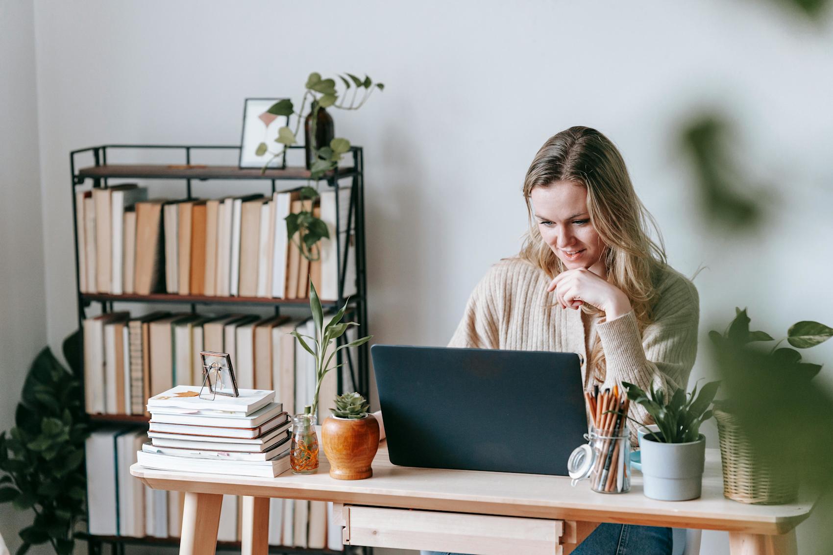 Bildet viser en blond kvinne som hviler haken i hånden, mens hun ser på en åpen laptop. Hun sitter på et kontor med grønne planter og er omringet av flere bøker.