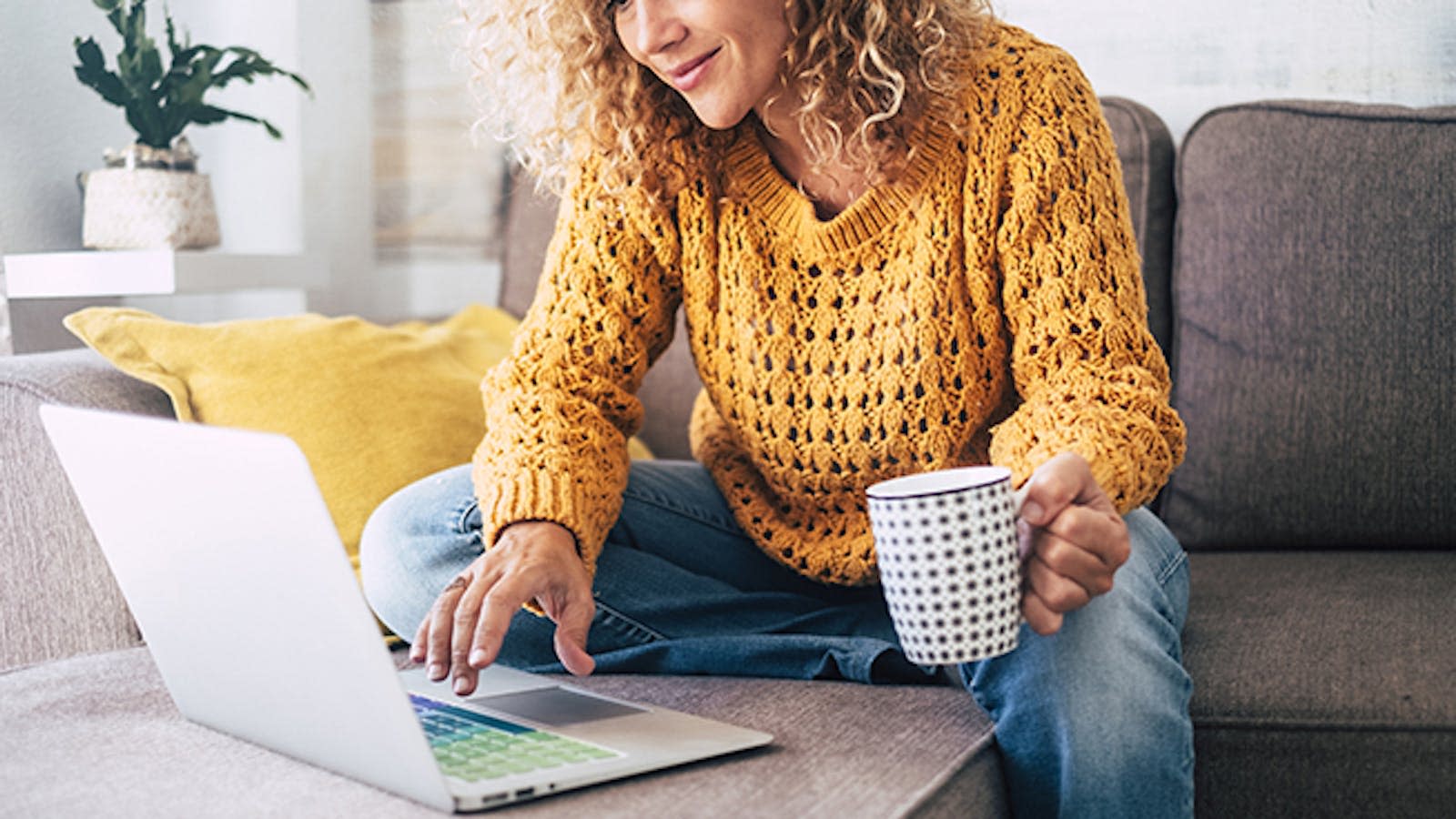 En dame sitter på sofaen og drikker te samtidig som hun er på en laptop