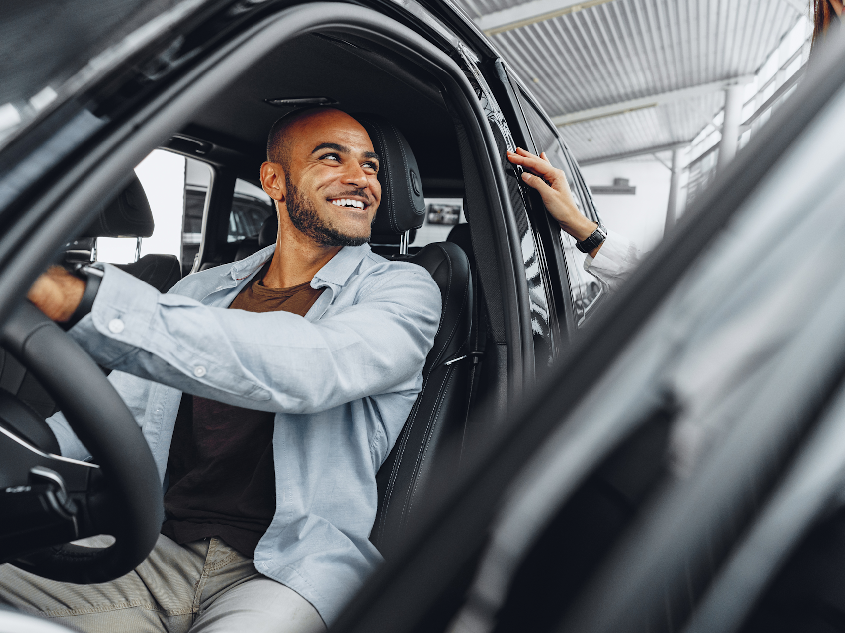 Smilende person i lyseblå skjorte sitter bak rattet i en bil i en bilforretning, mens en ansatt hjelper til.