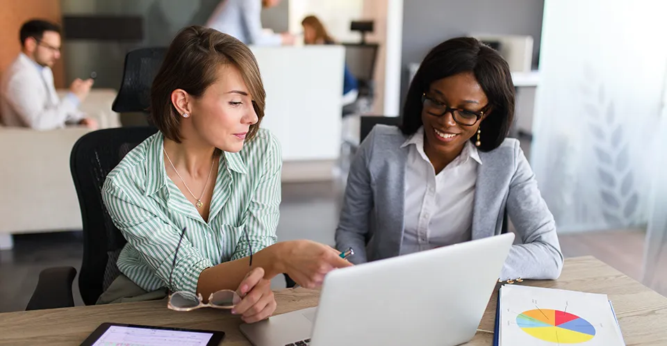 In an office, two people sit and talk at a desk. A laptop and a document displaying a pie chart sit on the table in front of them.