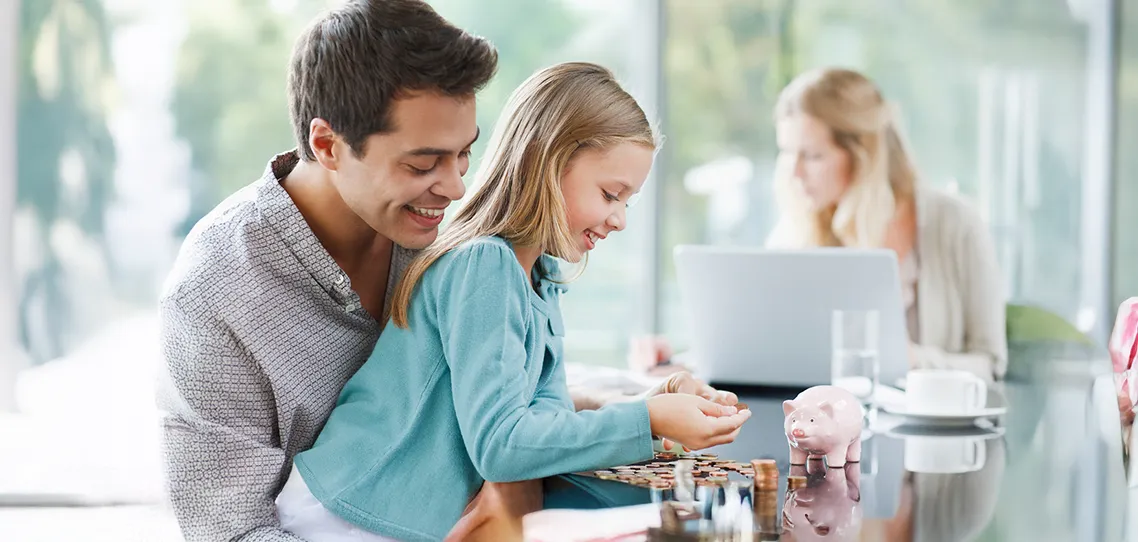 A couple and their little girl sit at a table in a brightly lit room. The father smiles as he watches his daughter carefully counting coins next to a pink piggy bank while the mother sits in front of a laptop in the background.